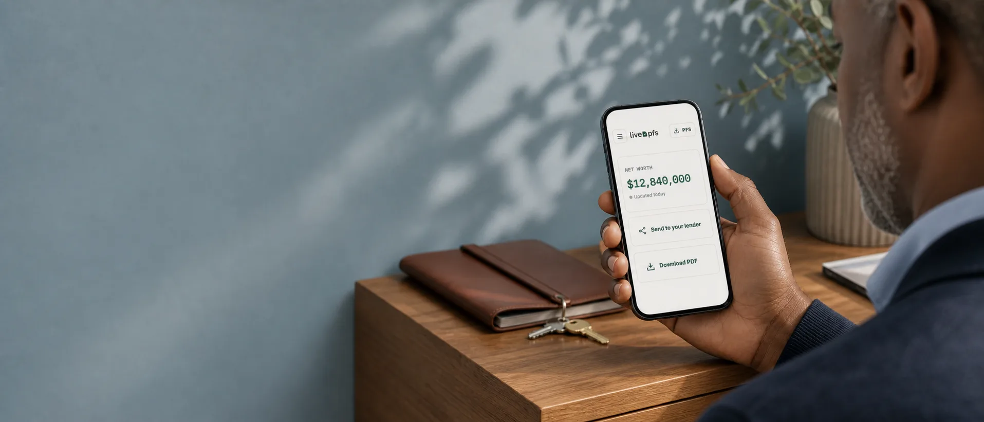 A borrower holds a phone showing an illustrative LivePFS net-worth summary — example figures, not customer data — at an oak side table against a slate-blue wall with soft leaf shadows
